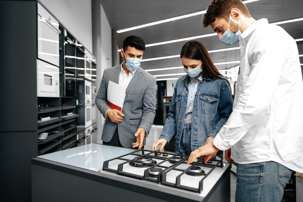 shop assistant hypermarket shows new model gas stove young couple all wearing medical masks Appliance Repair Appointment