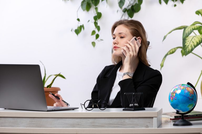 front view young beautiful lady white shirt black jacket using her laptop front table talking phone with leaves hanging 7 Essential TCPA Compliance Cold Calling Rules